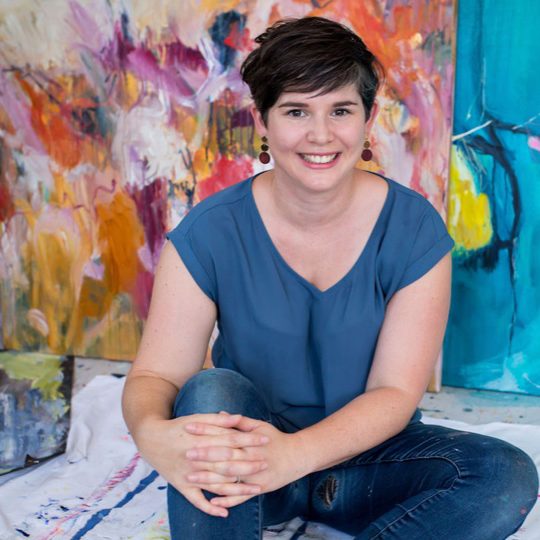 Alexandra Plim, artist sitting on studio floor on drop sheet, hands on knee, background includes large abstract, colourful art work.. Large paint brush strokes 
